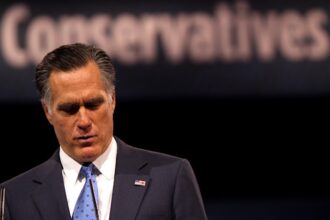 Image of Mitt Romney at a podium looking down with blurred sign in background that reads "Conservatives"