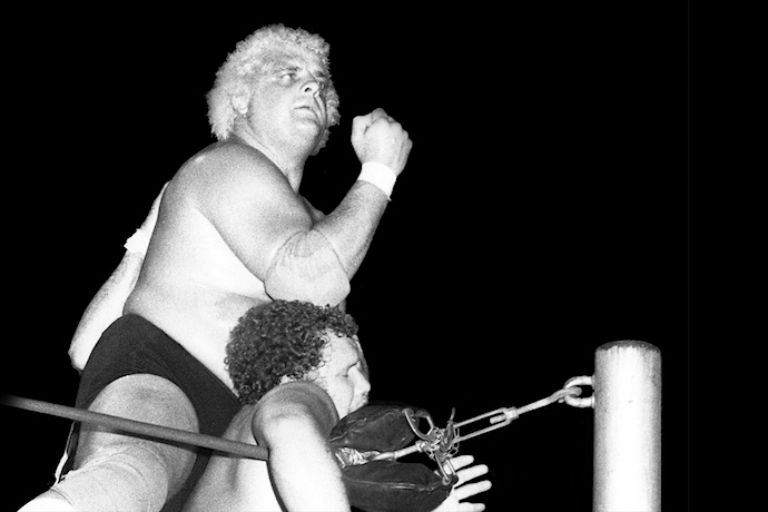 A blonde curly haired wrestler grinds an elbow into another who is hanging listlessly on the ropes.